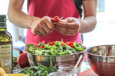 Hände einer erwachsenen Person, die Tomaten schneidet, auf dem Tisch davor eine Schüssel mit grünem Salat