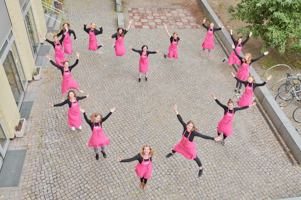 Gruppenfoto von Mitarbeitenden von RESTLOS GLÜCKLICH e. V.: 16 Personen tragen eine pinke Schürze mit RESTLOS GLÜCKLICH Logo, sind in Form eines Herz im Freien aufgestellt und springen in die Höhe