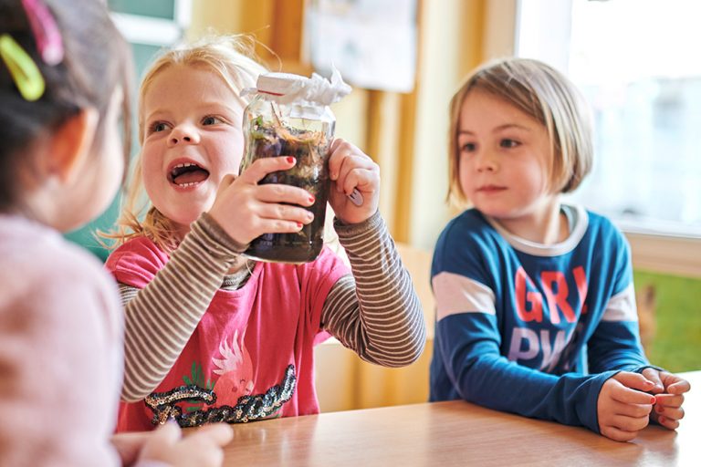 Drei Vorschulkinder, eins davon hält lachend ein selbstgebasteltes Kompostglas in beiden Händen