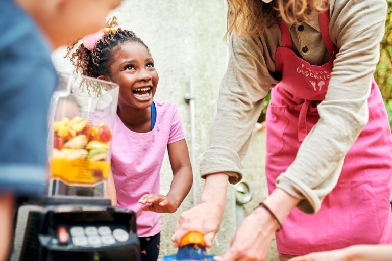 Ein lachendes Vorschulkind steht draußen neben einem Mixgerät gefüllt mit geschnittenem Obst. Daneben die Arme und Hände einer mitarbeitenden Person von RESTLOS GLÜCKLICH e. V., die eine Orange auspressen.