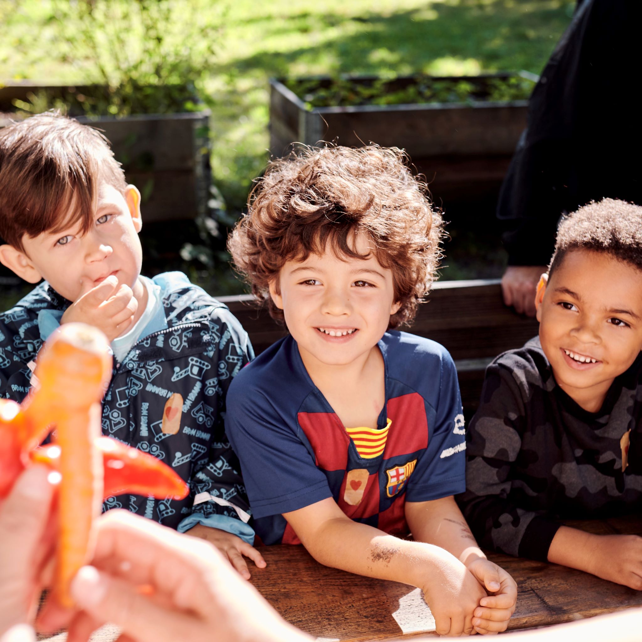 Drei Vorschulkinder schauen sich eine zweibeinige Karotte und eine Paprika an, die zwei Hände einer erwachsenen Person halten