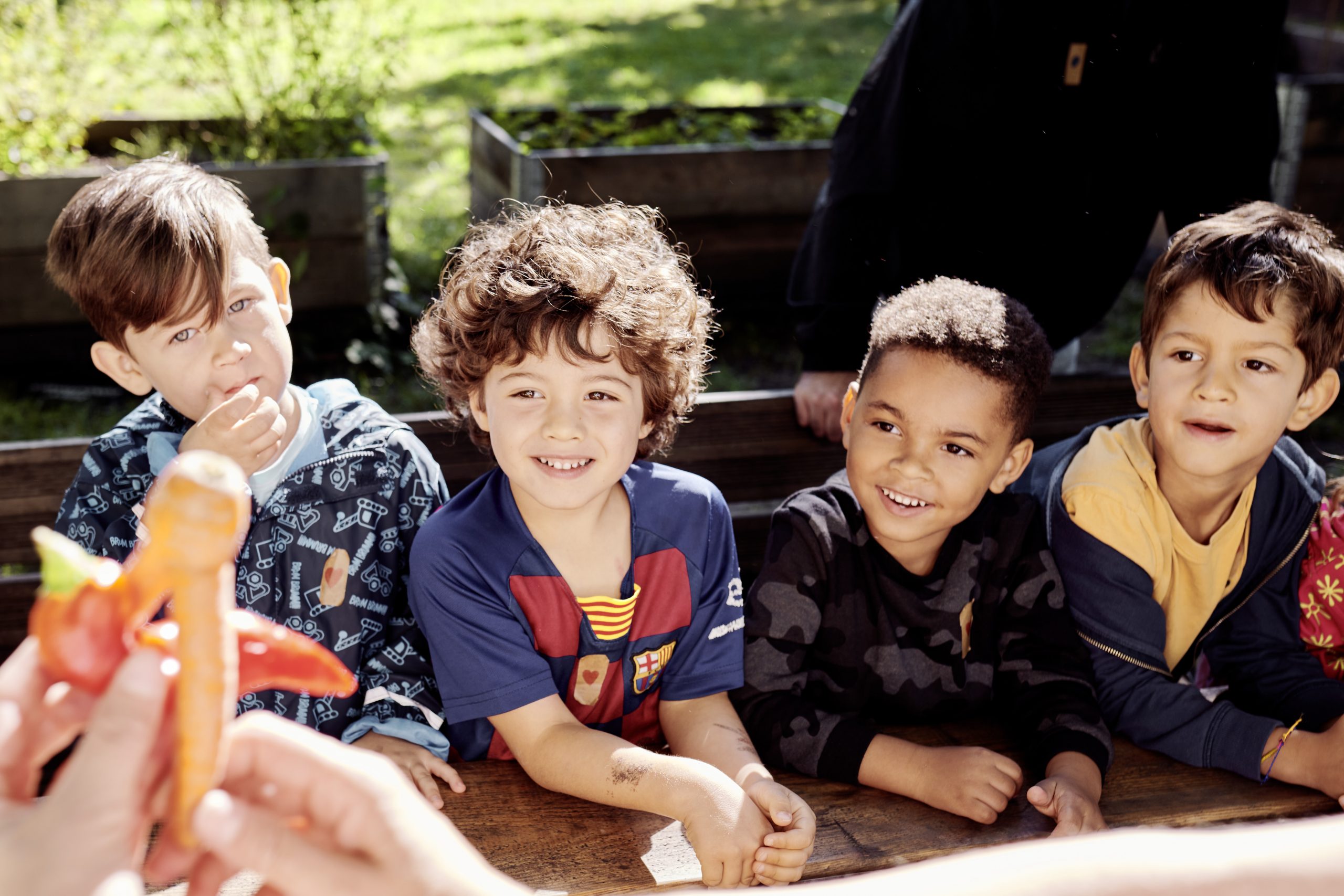 Vier Vorschulkinder betrachten lächelnd krummes und gerettetes Gemüse in den Händen einer mitarbeitenden Person von RESTLOS GLÜCKLICH e. V.