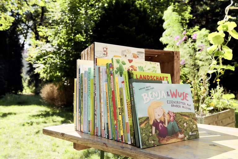 Mehrere Kinderbücher rund um nachhaltige Ernährung stehen auf einem Tisch. Beim vordersten Buch ist die Titelseite zu sehen: „Benja & Wuse. Essensretter auf großer Mission."