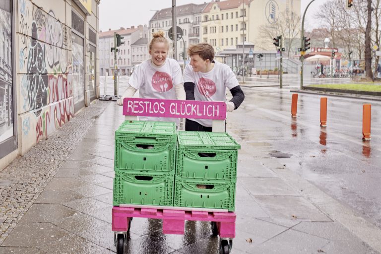 Zwei mitarbeitende Personen von RESTLOS GLÜCKLICH schieben lachend eine Europalette auf Rollen auf einem Gehsteig. Auf der Europalette sind 4 Kisten mit geretteten Lebensmitteln. Dahinter die Aufschrift „RESTLOS GLÜCKLICH".