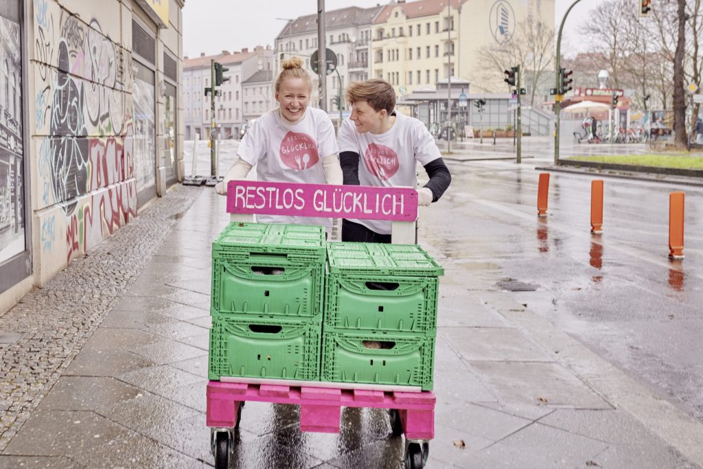 Zwei mitarbeitende Personen von RESTLOS GLÜCKLICH schieben lachend eine Europalette auf Rollen auf einem Gehsteig. Auf der Europalette sind 4 Kisten mit geretteten Lebensmitteln. Dahinter die Aufschrift „RESTLOS GLÜCKLICH".
