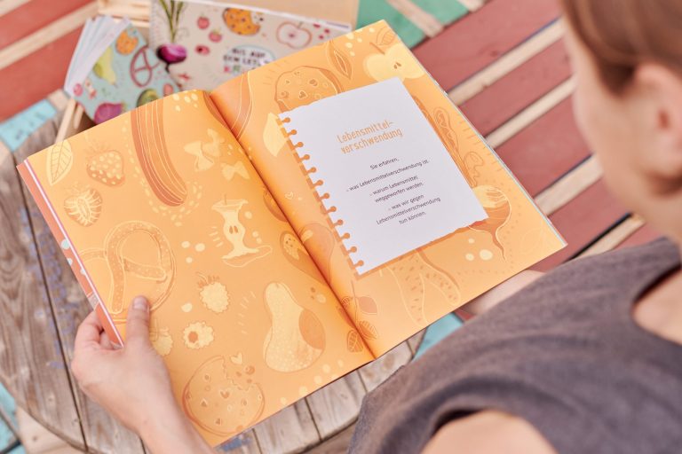 Die Händer einer Person halten das aufgeschlagene Handbuch des Kitaprojekts „Bis auf den letzten Krümel" von RESTLOS GLÜCKLICH e. V.