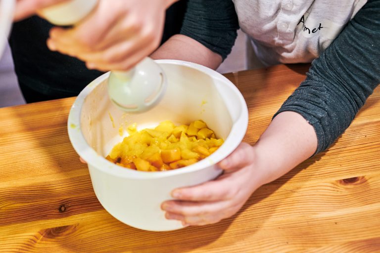 Zwei Kinderhände halten eine Schüssel, zwei andere Kinderhände halten einen Pürierstab und pürieren den Inhalt der Schüssel