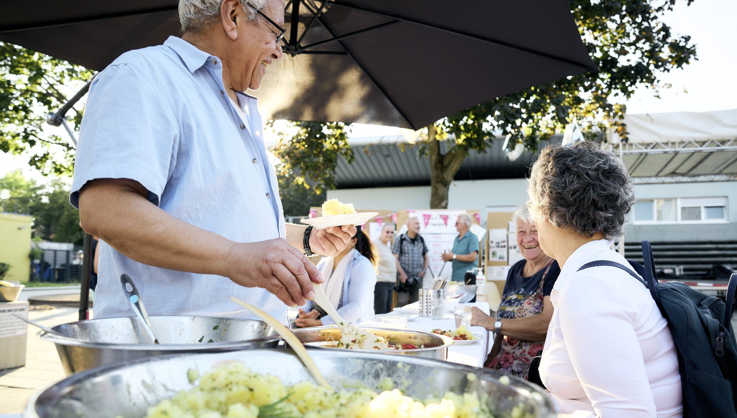 Teilnehmende eines Workshops im Freien lachen miteinander. Eine Person bedient sich ein einem Buffet.