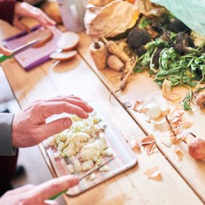 Die Hände von zwei erwachsenen Personen schneiden Brot und Zwiebeln auf Schneidebrettern an einem Holztisch.