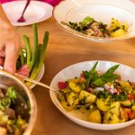 Eine Hand löffelt sich von einem Buffet-Tisch etwas auf den eigenen Teller.