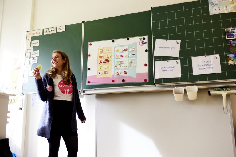 Nina Carryer von RESTLOS GLÜCKLICH e. V. steht lachend vor dem Kühlschrankspielposter, das an einer Tafel in einem Klassenzimmer hängt