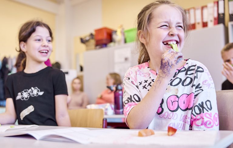 Zwei Grundschulkinder sitzen lachend an einem Tisch im Klassenzimmer. Das Kind auf der rechten Seite beißt in ein Gemüse.
