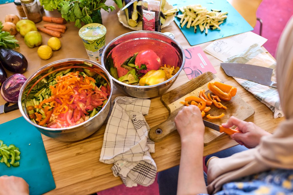 Wir schauen einer erwachsenen Frau über die Schulter, wie sie gerade an einem Tisch sitzt und verschiedenes Gemüse, wie Karotten und Paprika für einen Koch-Workshop schneidet.