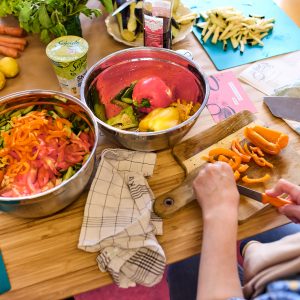Wir schauen einer erwachsenen Frau über die Schulter, wie sie gerade an einem Tisch sitzt und verschiedenes Gemüse, wie Karotten und Paprika für einen Koch-Workshop schneidet.
