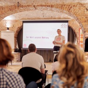 Edith Timm von RESTLOS GLÜCKLICH e. V. steht vor einer Gruppe Erwachsener und hält einen Vortrag.