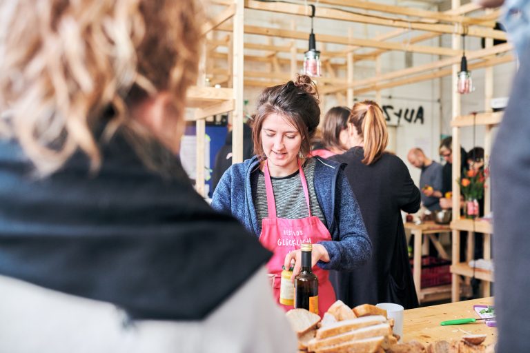 Ann-Kathrin Ridder von RESTLOS GLÜCKLICH e. V. stellt ein Glas Senf auf einem Tisch in einer Küche ab. Um sie herum und im Hintergrund sind mehrere Teilnehmende des Koch-Workshops, die sich unterhalten und Lebensmittel schneiden.