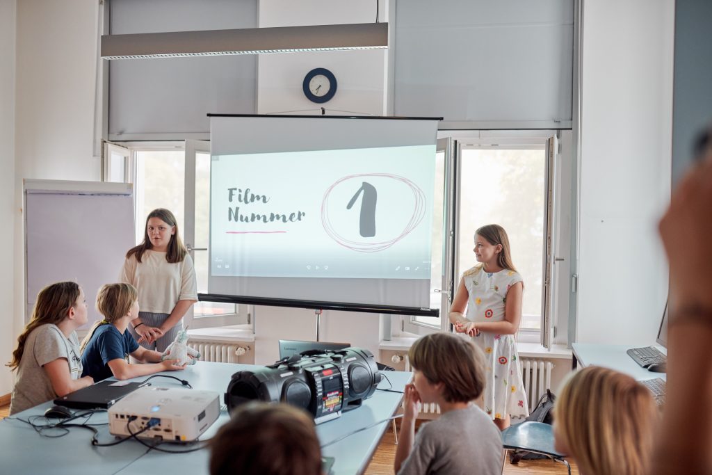 Zwei Grundschulkinder stehen vor einer Gruppe anderer Grundschulkinder und stellen auf einer Leinwand ihren Film aus einem stop-motion Projekt vor.