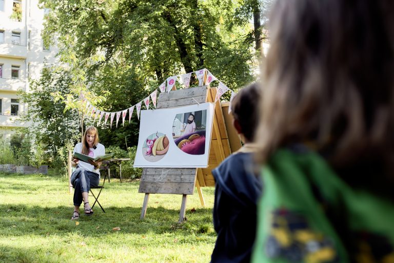 Wenke Heuts von RESTLOS GLÜCKLICH e. V. sitzt im freien auf einem Stuhl und liest einer Gruppe Vorschulkinder aus dem Kinderbuch "Benja und Wuse" vor