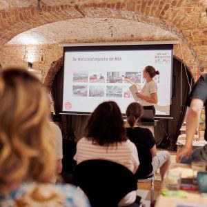 Edith Timm von RESTLOS GLÜCKLICH e. V. hält einen Vortrag. Mehrere Teilnehmende sitzen in Stuhlreihen vor Edith und hören zu.