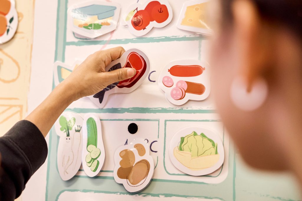 Beim Kühlschrankspiel: Eine Hand platziert die Illustration eines Stecks und eines Fischs in ein Fach des Kühlschrankposters