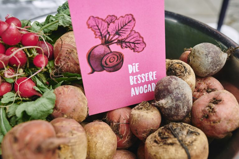 Eine Schüssel mit Roter Bete und einem Bund Radieschen. auf dem Gemüse liegt eine pinke Karte mit der Illustration einer Roten Bete und der Aufschrift „Die bessere Avocado"