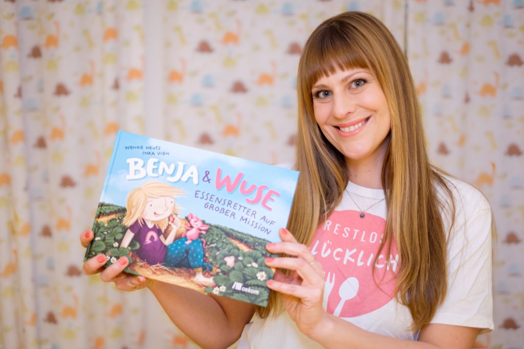 Wenke Heuts von RESTLOS GLÜCKLICH e. V. hält lächelnd das Kinderbuch „Benja & Wuse. Essensretter auf großer Mission" in der Hand.