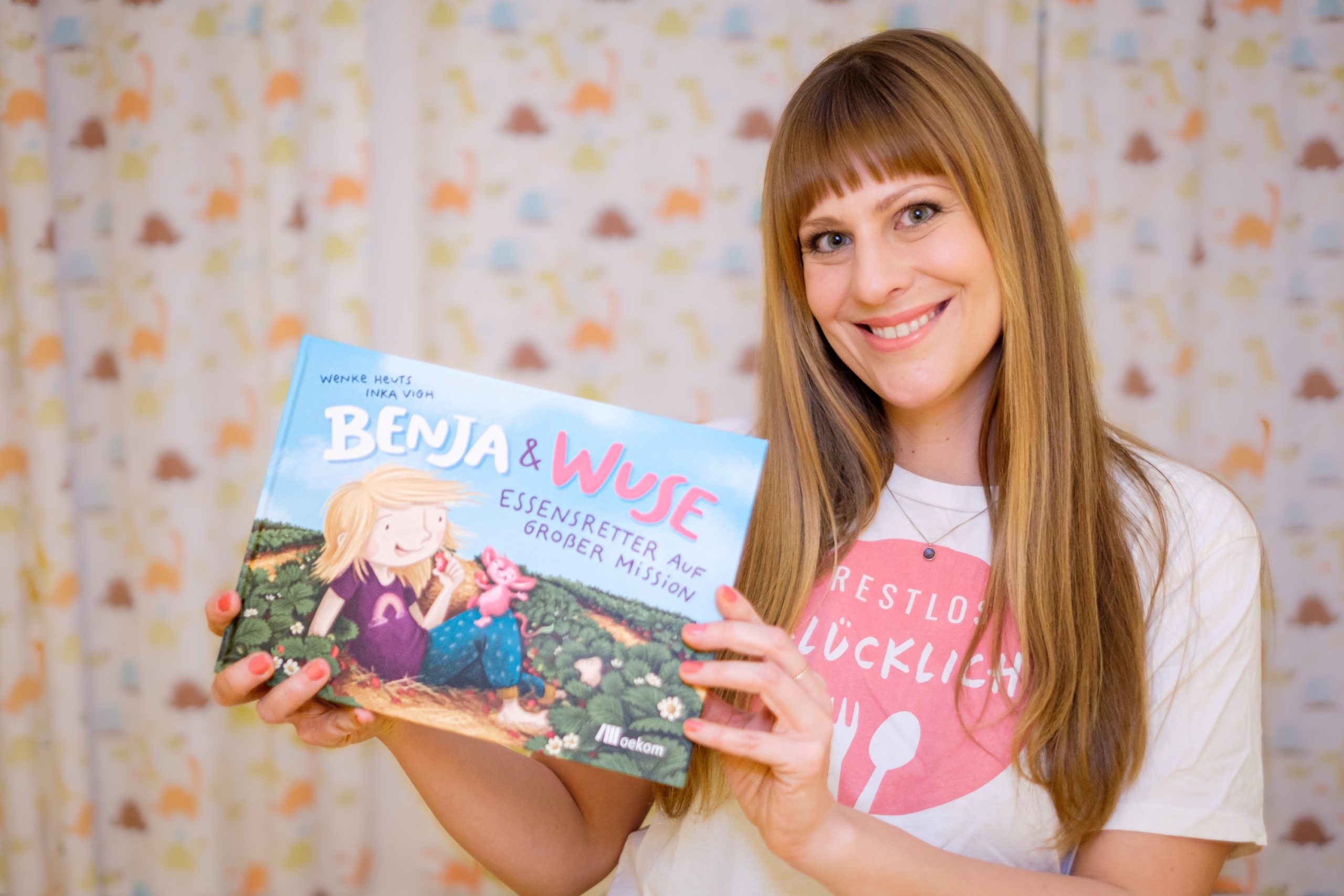 Wenke Heuts von RESTLOS GLÜCKLICH e. V. hält lächelnd das Kinderbuch „Benja & Wuse. Essensretter auf großer Mission" in der Hand.