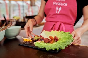 Snacks aus geretteten Lebensmitteln auf einem Schiefertablett auf einem Tresen, dahinter der Oberkörper einer Person mit pinker RESTLOS GLÜCKLICH Schürze