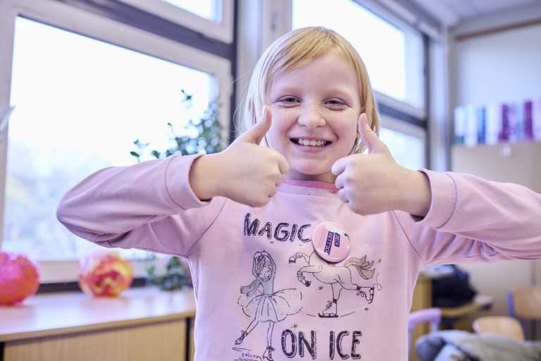 Ein Mädchen im Grundschulalter schaut freudestrahlend in die Kamera und hält beide Daumen nach oben.