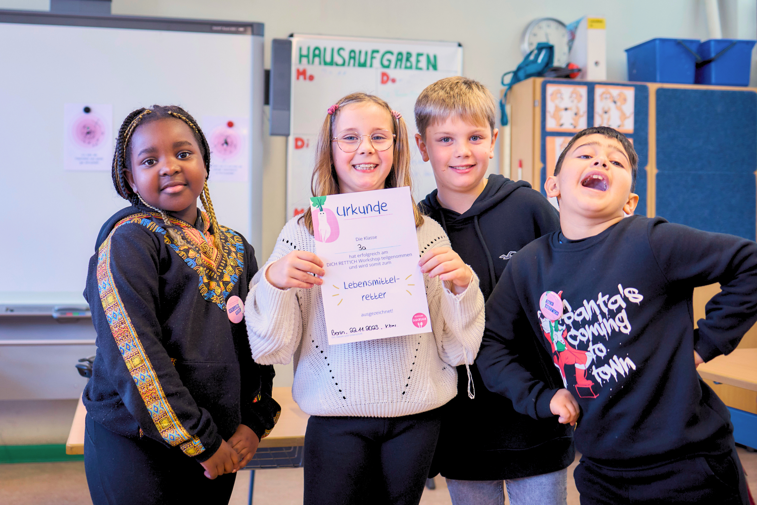 Zwei Mädchen und zwei Jungs im Grundschulalter stehen in ihrem Klassenzimmer und strahlen in die Kamera. Ein Mädchen hält stolz die Urkunde hoch, die jede Klasse am Ende des Workshoptages im Projekt "DICH RETT'ICH" zu Lebensmittelretter*innen auszeichnet.