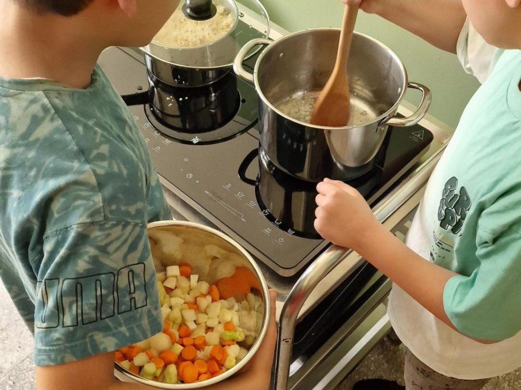 Zwei Jungs im Grundschulalter stehen am Kochfeld. Der eine schwitzt gerade die Zwiebeln im Kochtopf an, der andere hält eine Schüssel voller Gemüse bereit.