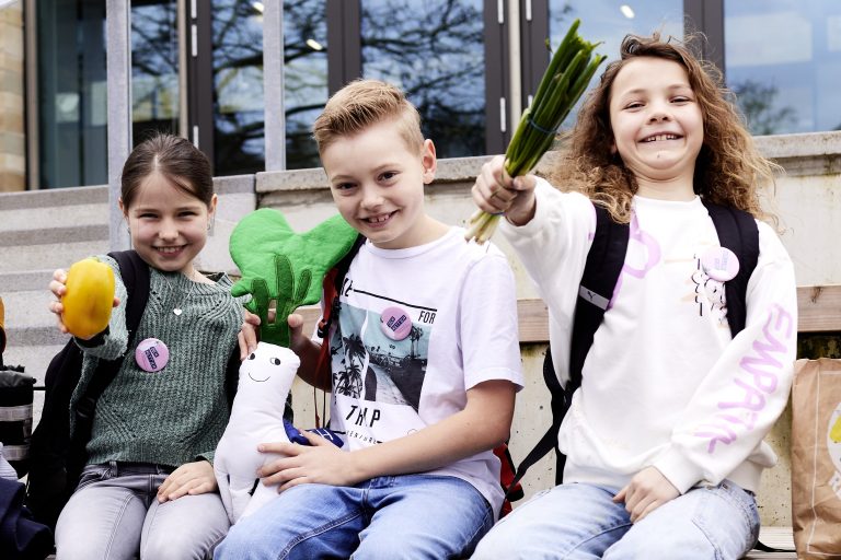 Drei Grundschulkinder sitzen lachend auf einer Treppe. Der Junge hält das Maskottchen Rubi Rett’ich in die Kamera, die Mädchen präsentieren eine Orange und eine Stange Porree.