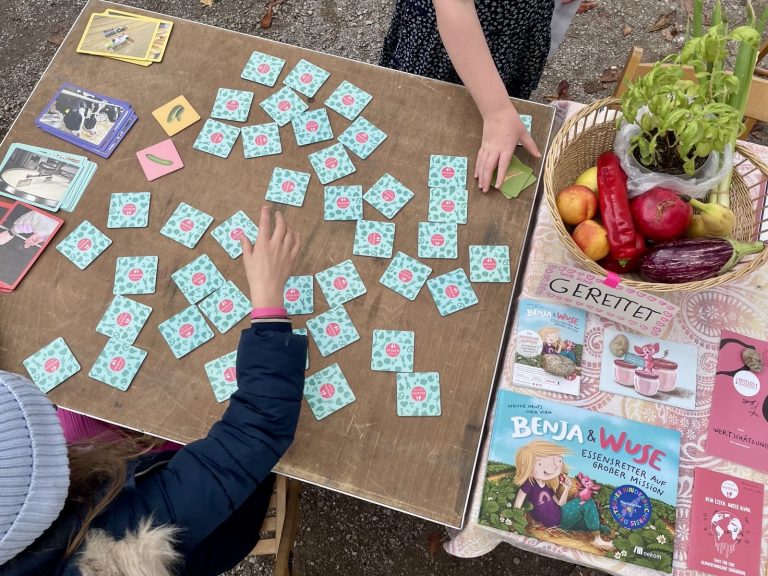 Das Memo-Spiel von RESTLOS GLÜCKLICH liegt auf einem Infostand-Tisch. Die Hände von zwei Kindern sind zu sehen, die das Spiel spielen.