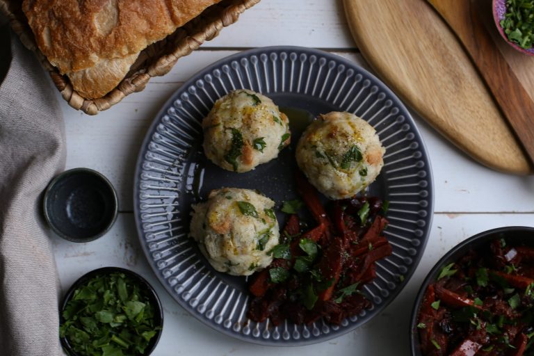 Ein taubenblauer Teller mit veganen Semmelknödeln und veganem Wurzelgemüse. Drumherum im Anschnitt zu sehen sind ein Korb mit Brot und kleine Schalen mit geschnittenen Kräutern.
