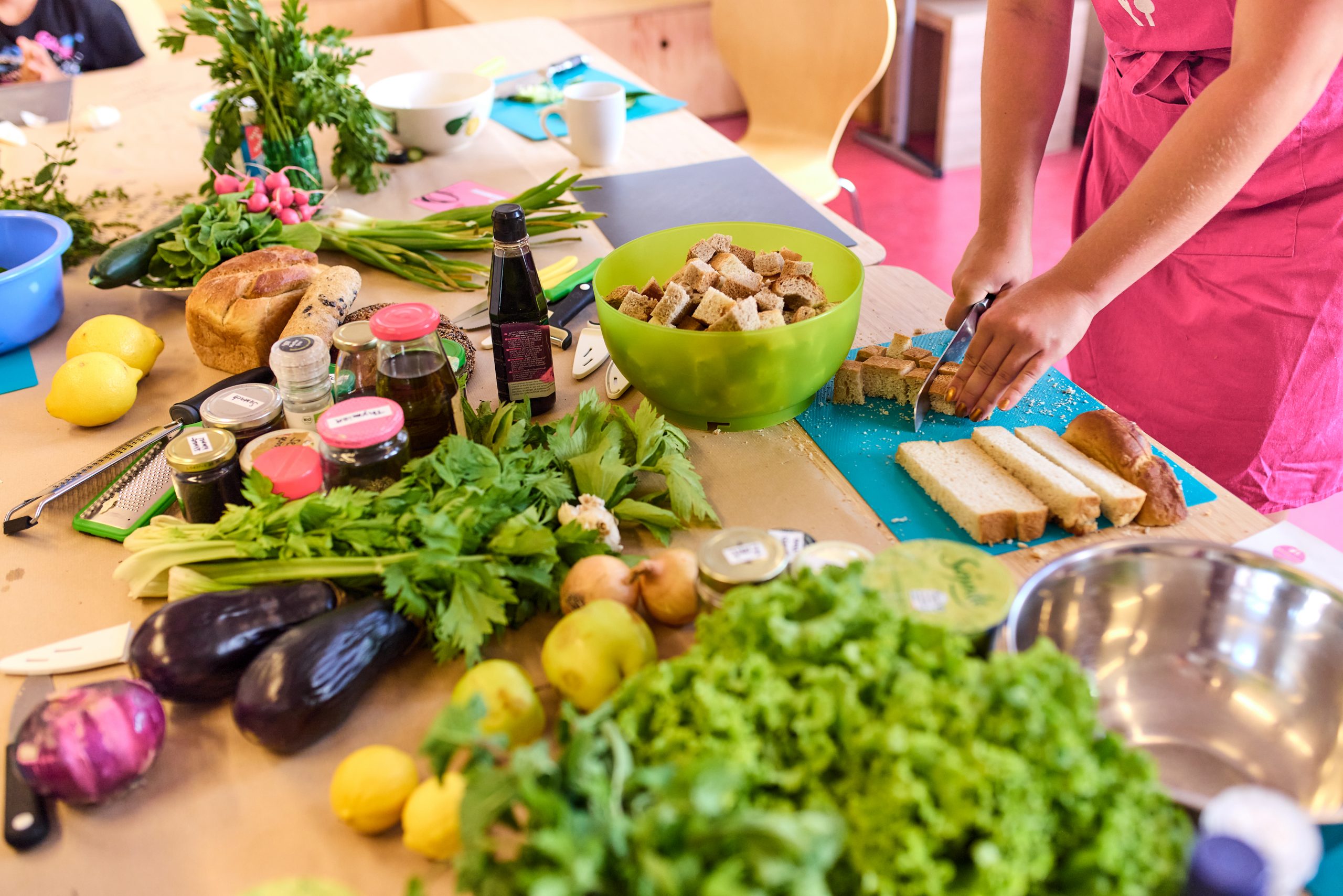 Ein gedeckter Tisch mit vielen Lebensmittel, darunter Salat, Zitronen und Auberginen. Zu sehen ist, wie eine Person in pinker Kochschürze gerade Brotscheiben in kleine Würfel für den Koch-Workshop vorbereitet.