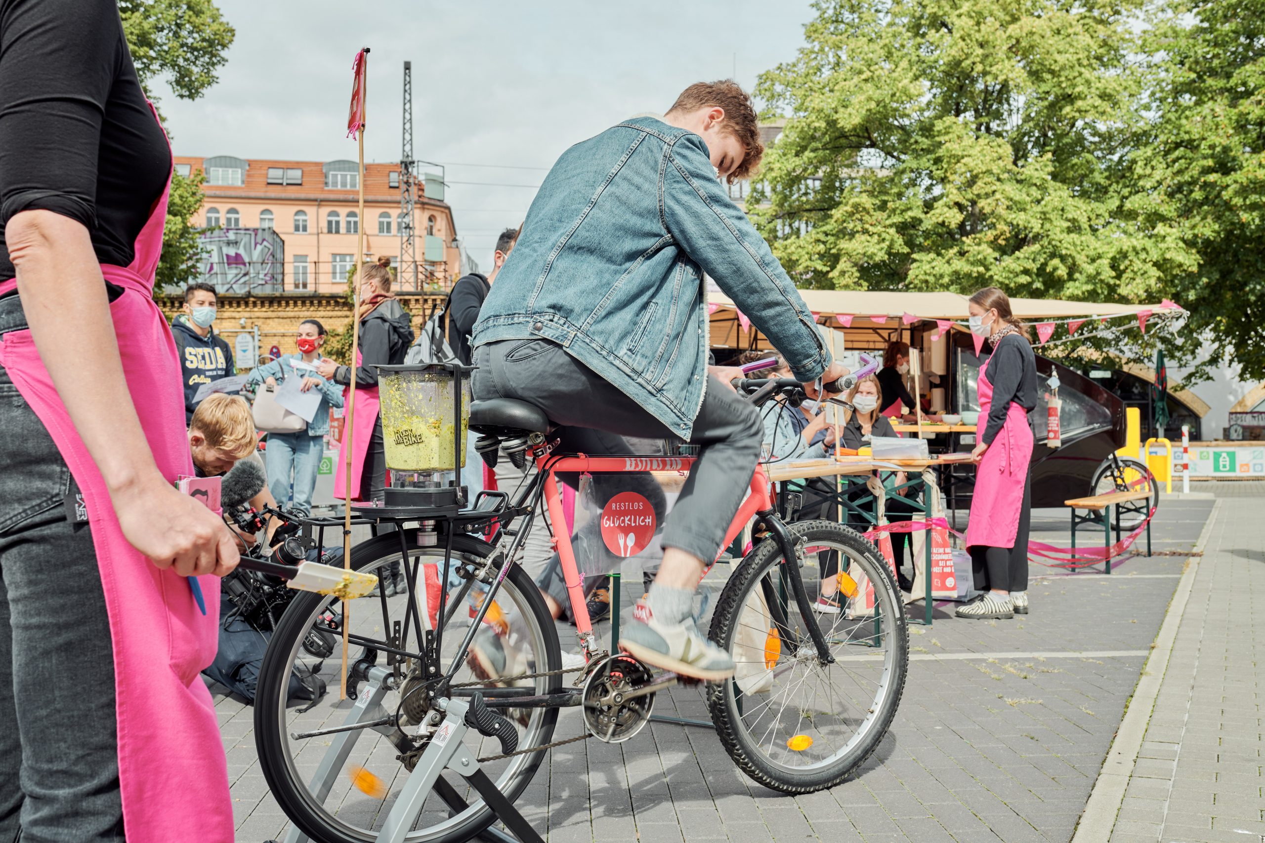 Bei einem Aktionsstand im Freien: Eine jugendliche Person sitzt auf dem Smoothiebike und tritt in die Pedale. Im Hintergrund ist der belebte Aktionsstand zu sehen.