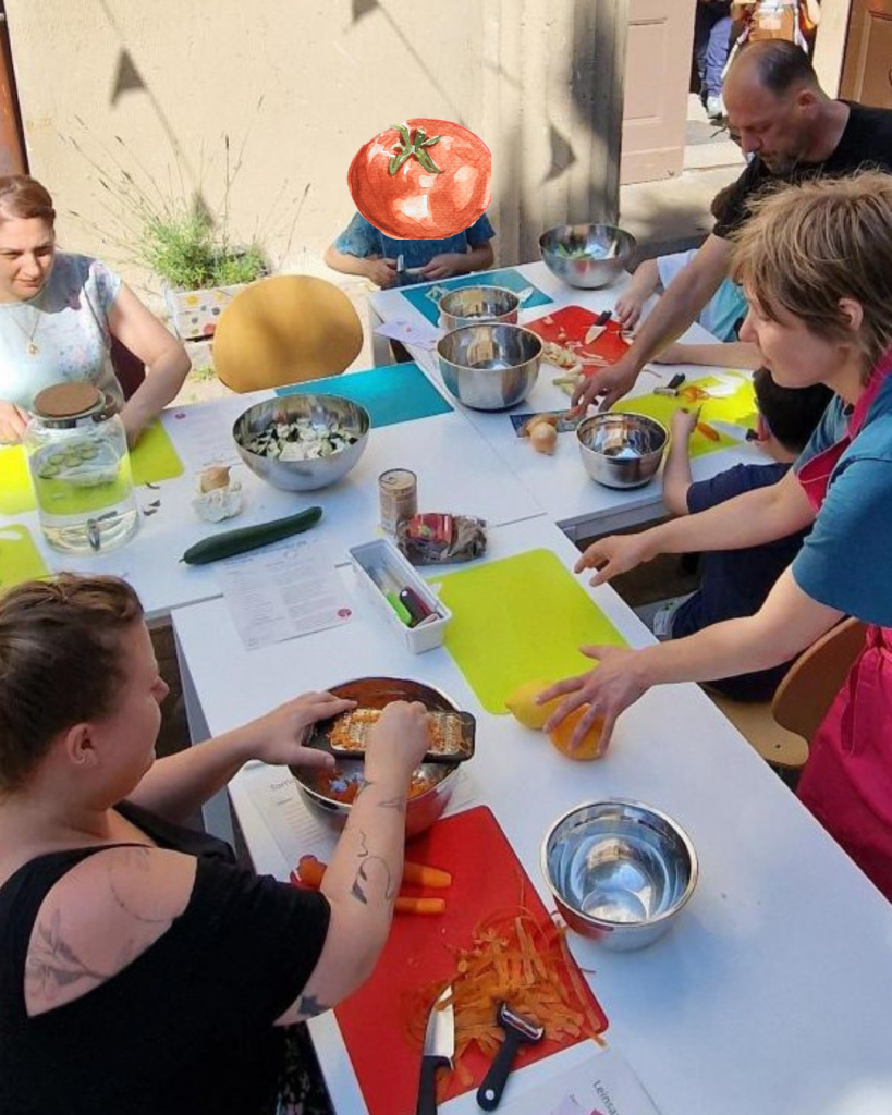 Erwachsene und Kinder sitzen draußen bei Sonnenschein an einer Tischtafel und schneiden Gemüse für den gemeinsamen Familien-Koch-Workshop.