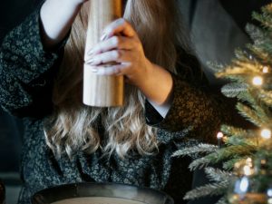 Die Hände einer Person halten eine Pfeffermühle über einen Teller und malen Pfeffer. Rechts am Bildrand ist ein Weihnachtsbaum zu sehen.