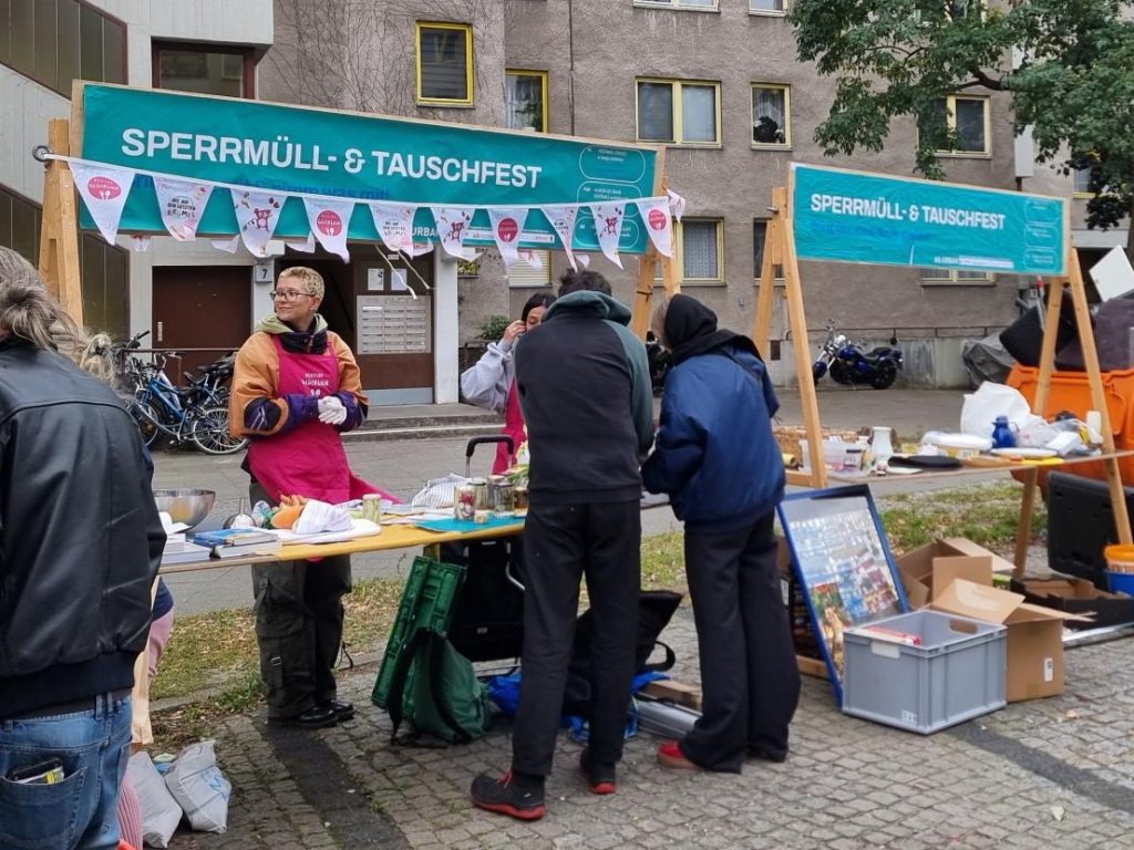 Zwei Erwachsene stehen draußen an einem Aktionsstand und sprechen mit dem Personal dahinter. Auf dem Stand ist das Schild „Sperrmüll- und Tauschfest“ zu sehen.