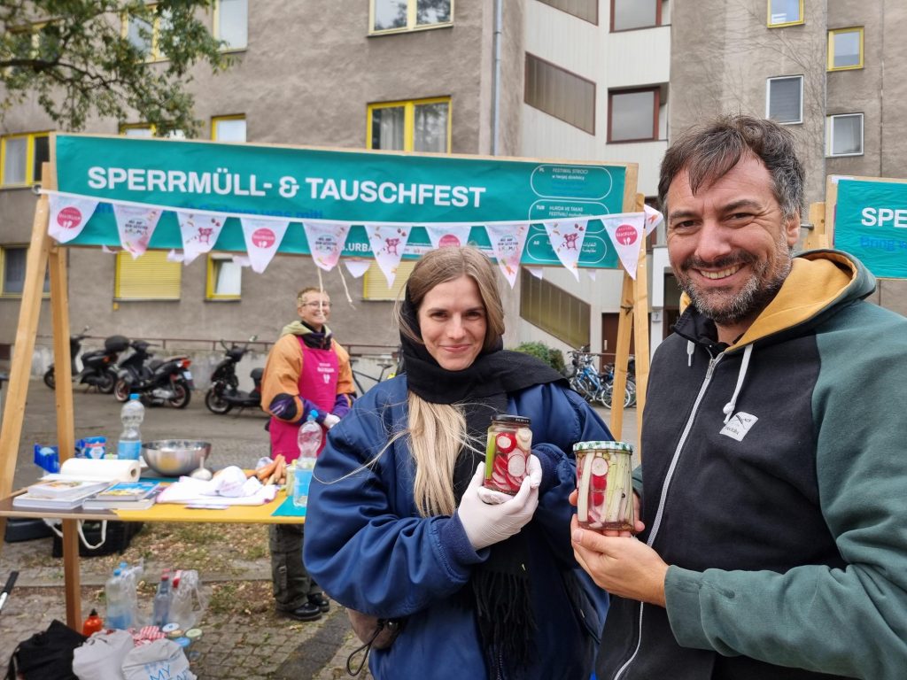 Eine Frau und ein Mann halten ein Glas mit fermentiertem Gemüse hoch und strahlen in die Kamera. Im Hintergrund steht eine Frau an einem Stand mit der Überschrift „Sperrmüll- und Tauschfest”.