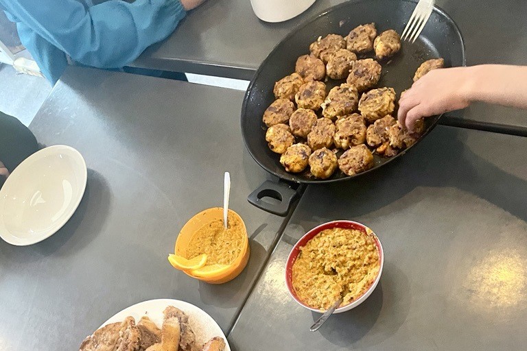 Blick von oben auf einen Tisch mit zwei kleinen Schüsseln Aufstrich und einer Pfanne mit Linsenbroletten. Am Rand sitzen Menschen.