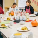 Zwei Frauen sitzen an einem gedeckten Tisch. In der Mitte stehen mehrere Flaschen Mineralwasser und drumherum Teller mit Suppe und Kürbispuffer sowie zwei Kürbisse.