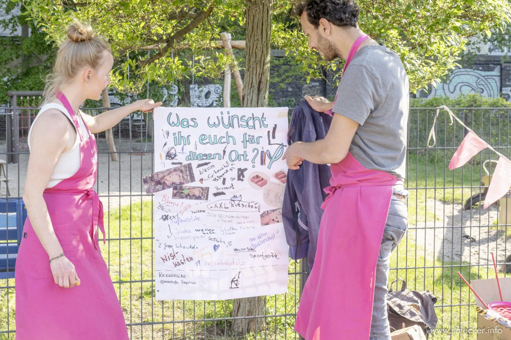 Eine Frau und ein Mann, beide in pinken Schürzen, stehen in einem Park und befestigen ein Plakat an einem Zaun mit der Aufschrift: "Was wünschst ihr euch für diesen Ort?"