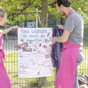 Eine Frau und ein Mann, beide in pinken Schürzen, stehen in einem Park und befestigen ein Plakat an einem Zaun mit der Aufschrift: "Was wünschst ihr euch für diesen Ort?"