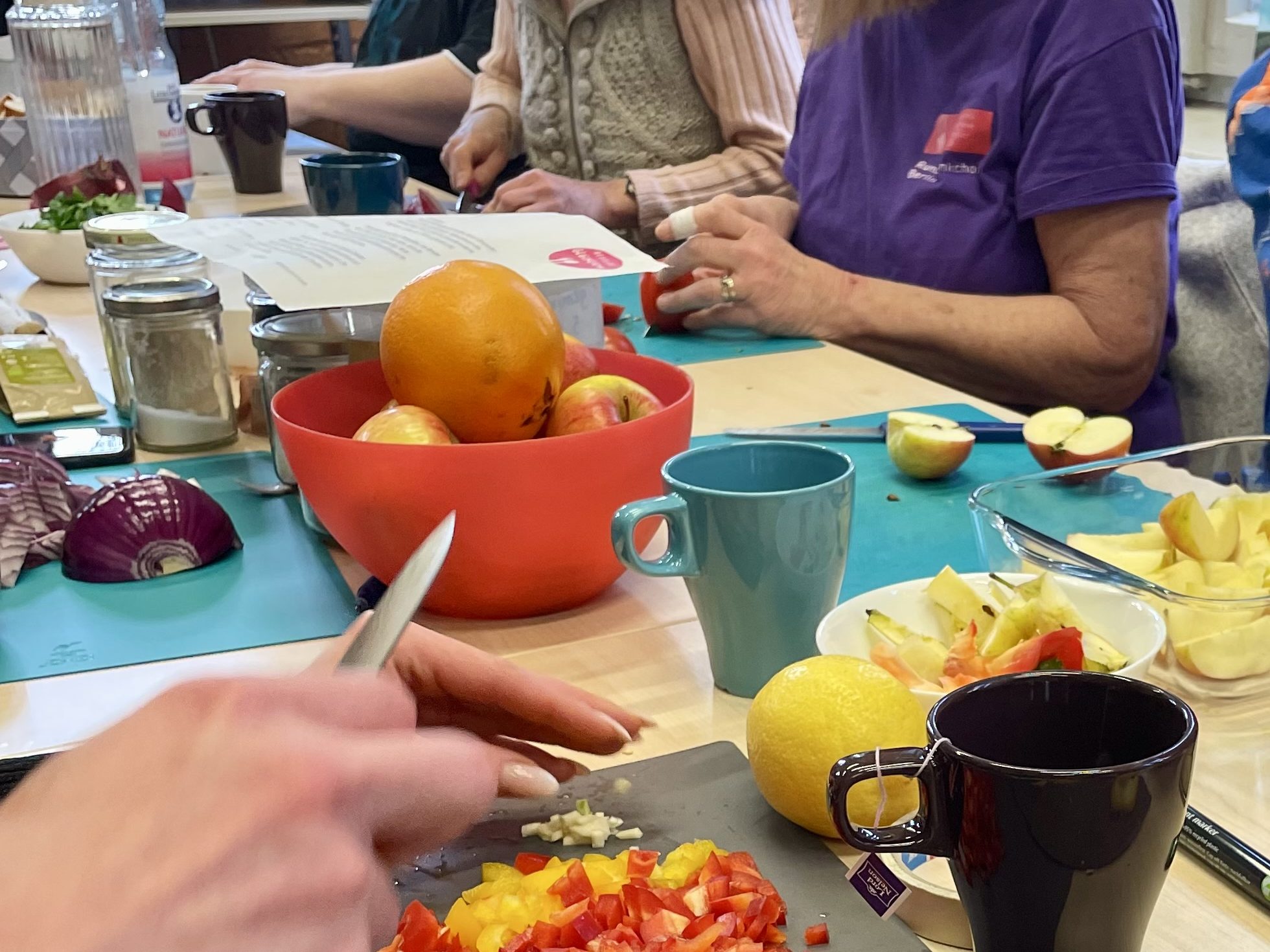Mehrere Erwachsene sitzen an einem Tisch und bereiten Obst und Gemüse zu, mit denen gekocht wird.