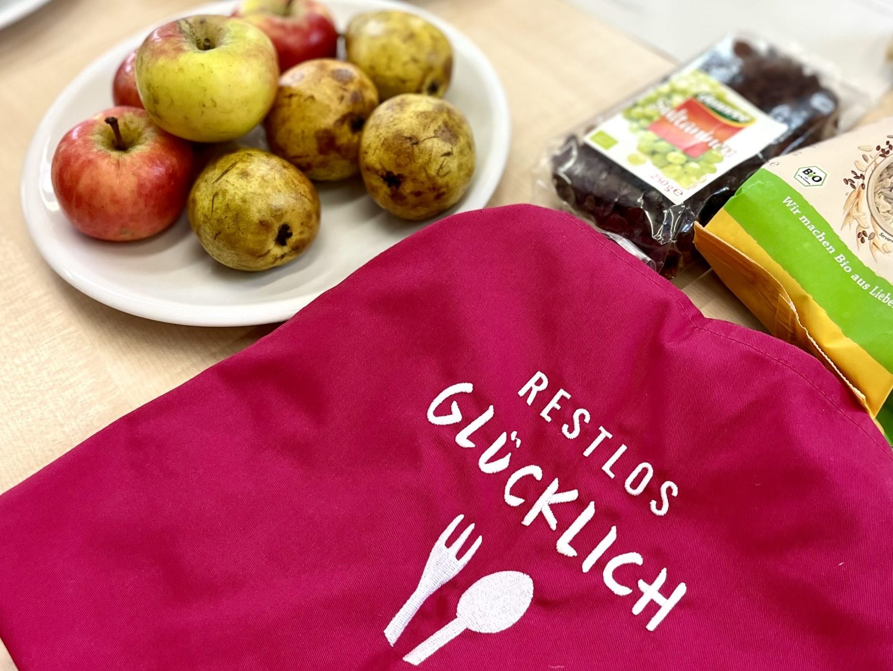 Ein Teller mit Äpfeln und Birnen liegt neben einer pinken Schürze mit der Aufschrift „Restlos Glücklich“ auf einem Tisch.