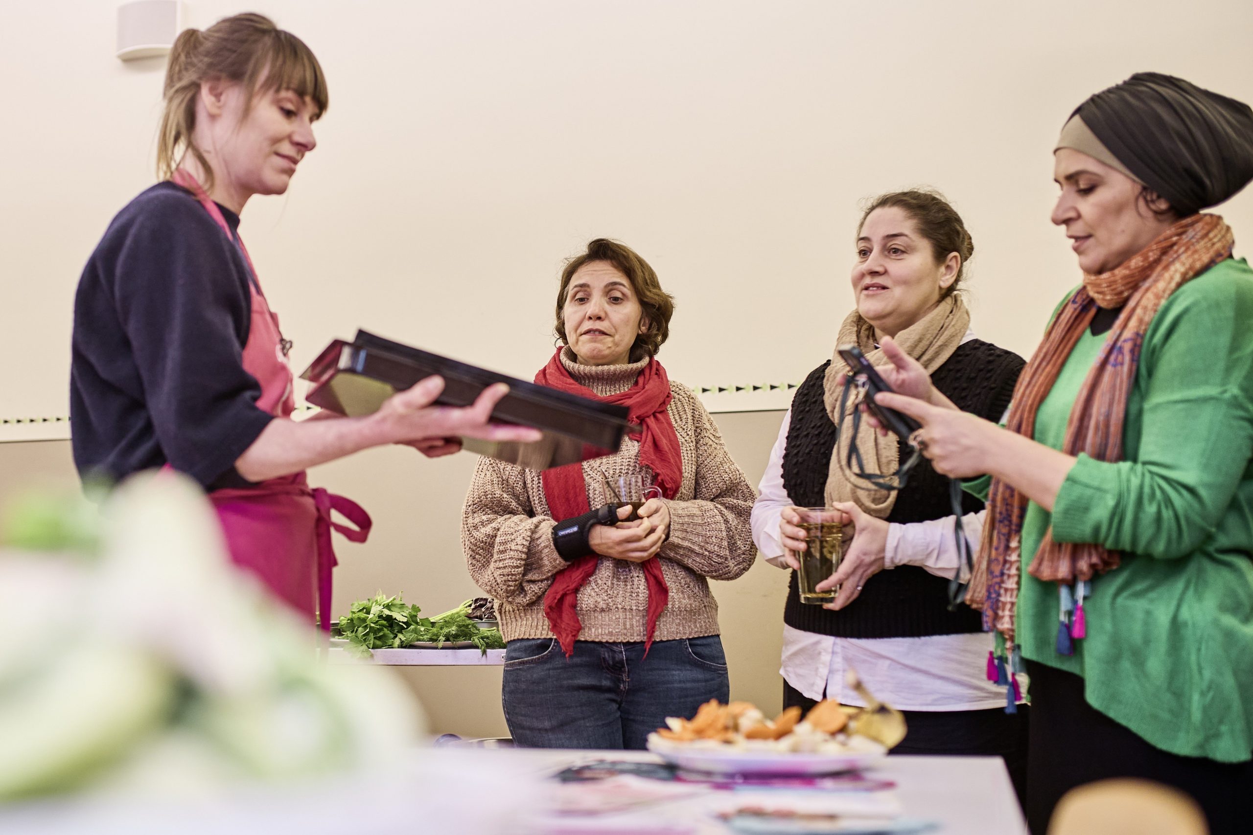 Eine Frau zeigt drei anderen Frauen ein Blech mit buntem Ofengemüse. Die Gruppe unterhält sich interessiert und macht Fotos vom Blech.