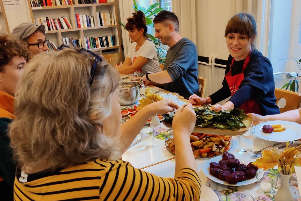 Sechs Erwachsene sitzen an einem Tisch und füllen ihre Teller mit Speisen, die sie zuvor im Koch-Workshop aus geretteten Lebensmitteln selbst zubereitet haben
