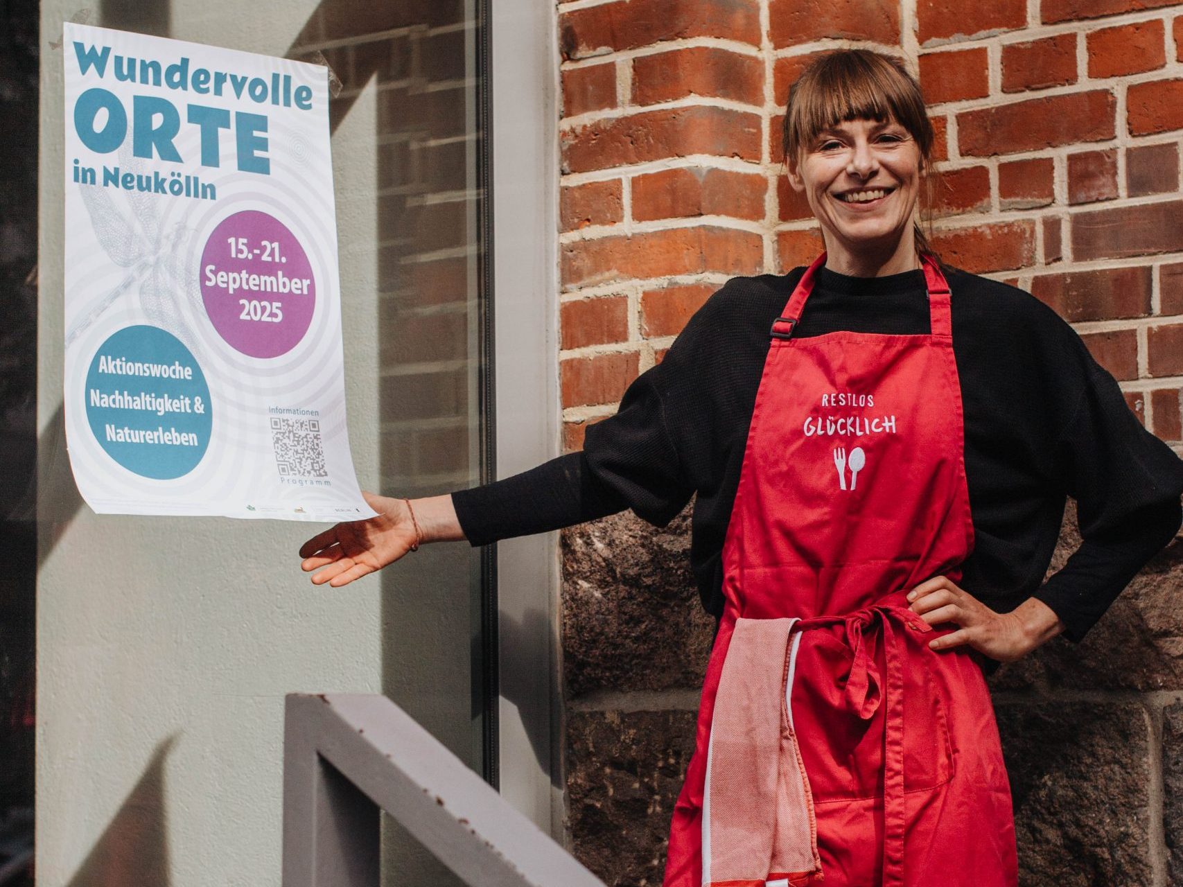 Ramona Holzer vom Projekt „Unsere Küche im Donaukiez“ steht lächelnd mit pinker Schürze vor einer Tür und zeigt auf ein Plakat der Aktionswoche „Wundervolle Orte“