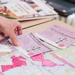 Auf einem Tisch liegen eine Stadtkarte und mehrere Postkarten mit der Aufschrift „Die Berliner Ernährungscoaches“. Eine Hand greift nach einer Postkarte.